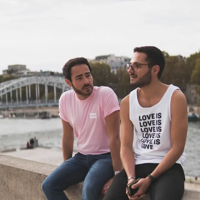 Zwei Personen, die an einem Fluss auf einer Mauer sitzen. Auf dem Oberteil einer Person steht &ldquo;Love is&rdquo;.